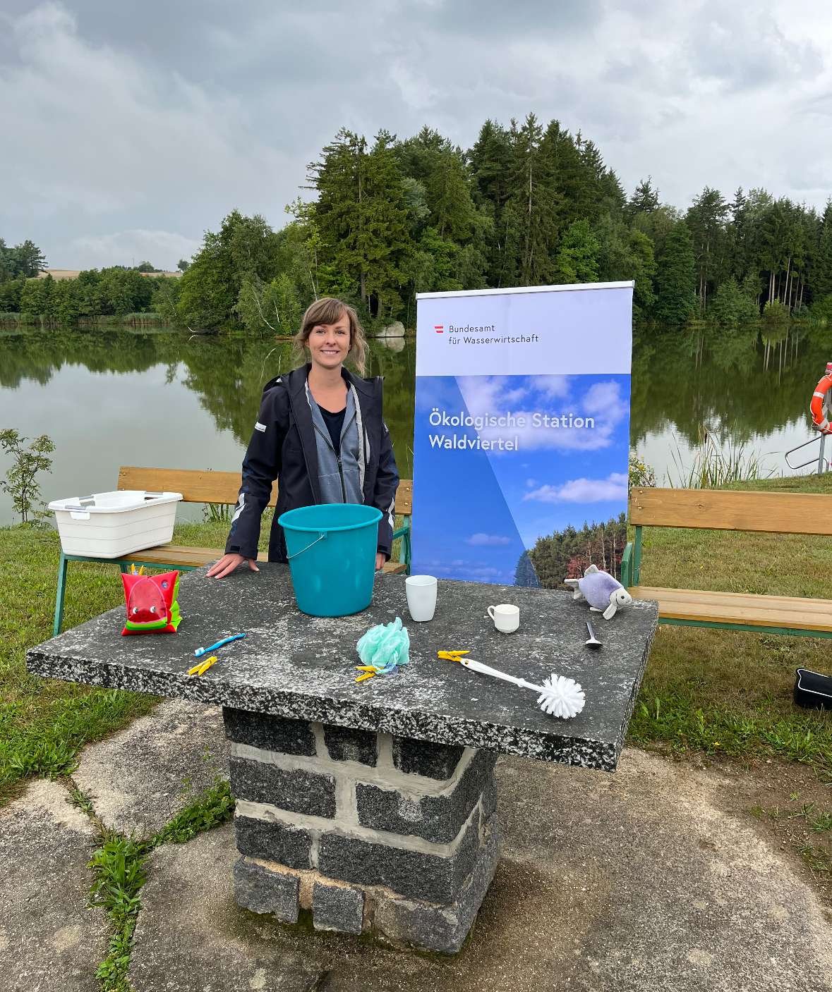 Elisabeth Peham hat einige Utensilien vorbereitet, um den Kindern ein spannendes Programm anzubieten. Elisabeth steht vor dem Teich an einem Steintisch mit den mitgebrachten Utensilien darauf.