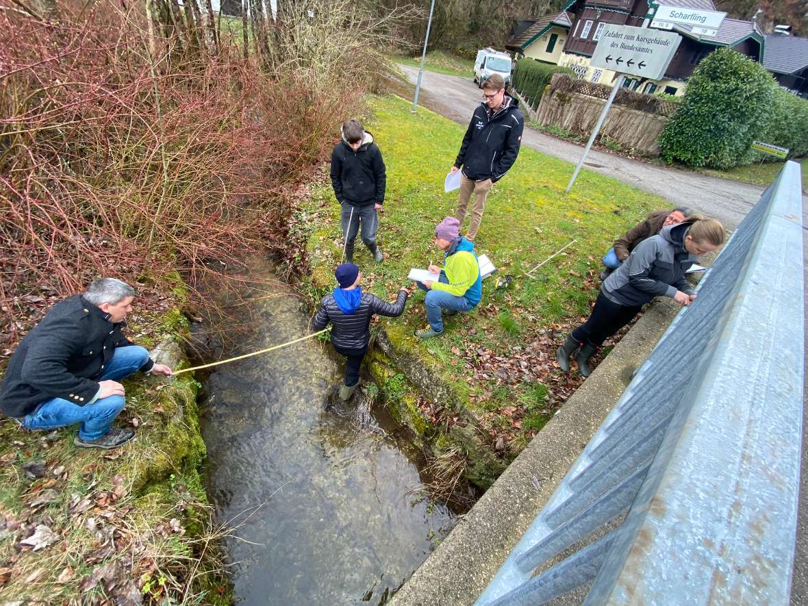 Die Teilnehmenden stehen bei einem Abluss vom See und Vermessung sowie Berechnung anhand eines Maßstabes den Abluss vor und nach einer Wasserentnahme sowie der Restwasserstrecke