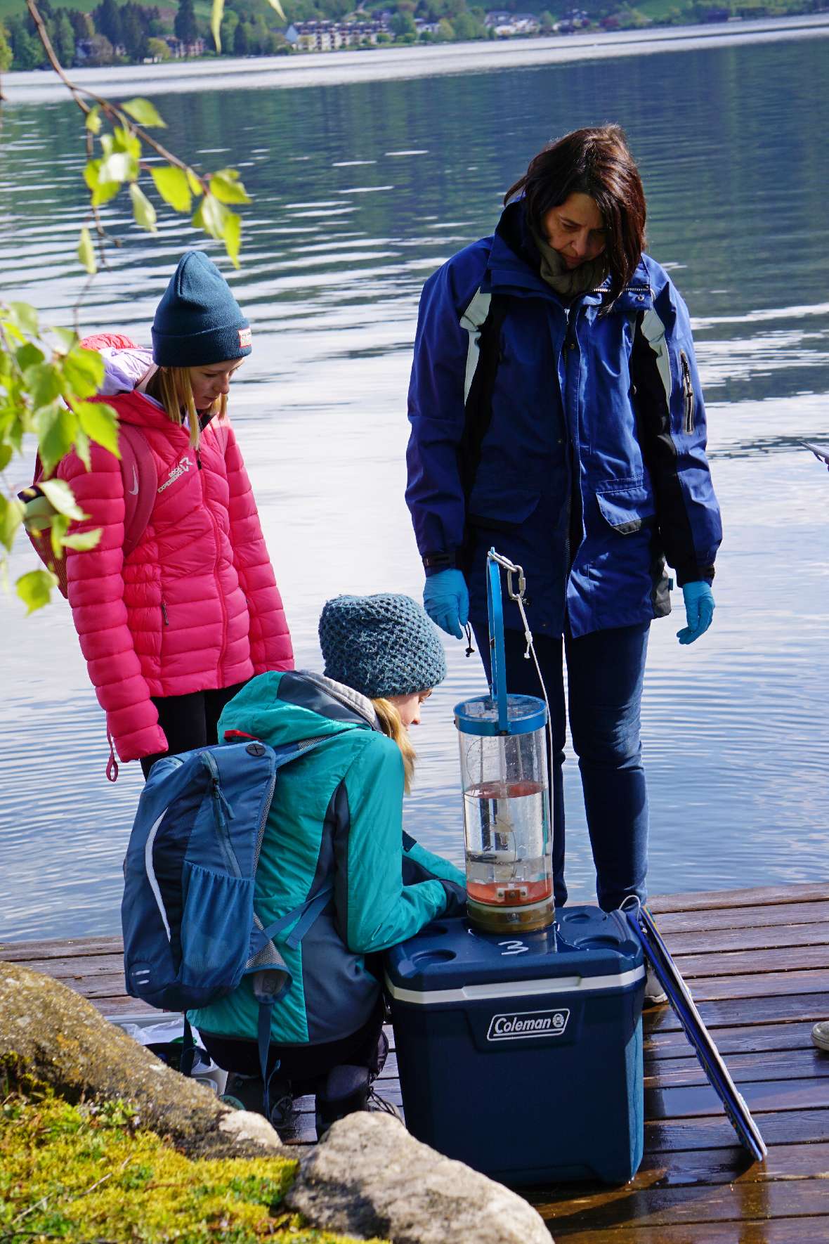 Abfüllen der mittels Schöpfer entnommenen Wasserprobe durch die zwei Teilnehmerinnen am Steg in Scharfling. Zu sehen sind zwei Mädchen und eine Mitarbeiterin des BAW sowie eine blaue Box und darauf ein Glasgefäß mit Wasser.
