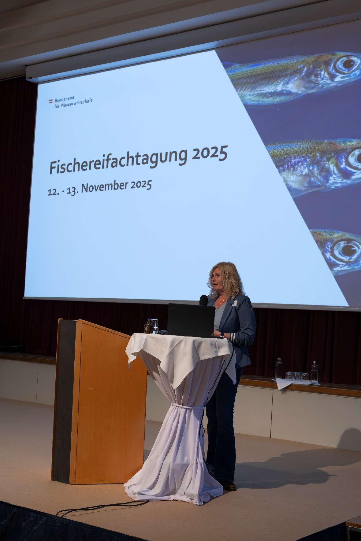Eröffnung der Tagung durch Gisela Ofenböck (BMLUK). Im Hintergrund sieht man eine Leinwand mit dem Text 'Fischereifachtagung 2025 12. - 13. November 2025' und eine Fischabbildungen.