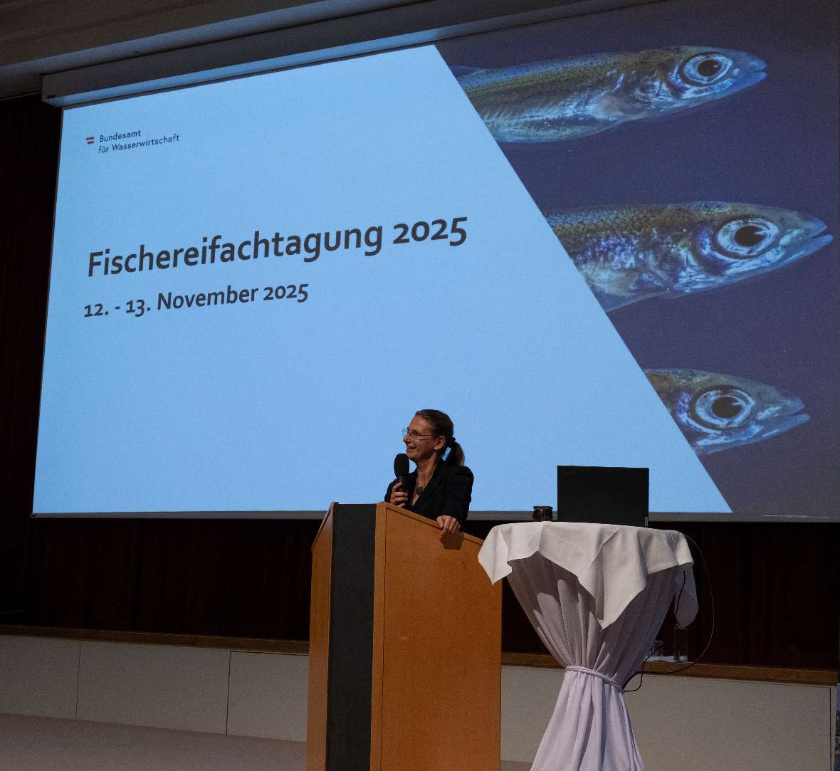Eröffnungsansprache durch Institutsleiterin Daniela Achleitern. Im Hintergrund sieht man eine Leinwand mit dem Text 'Fischereifachtagung 2025 12. - 13. November 2025' und eine Fischabbildungen.