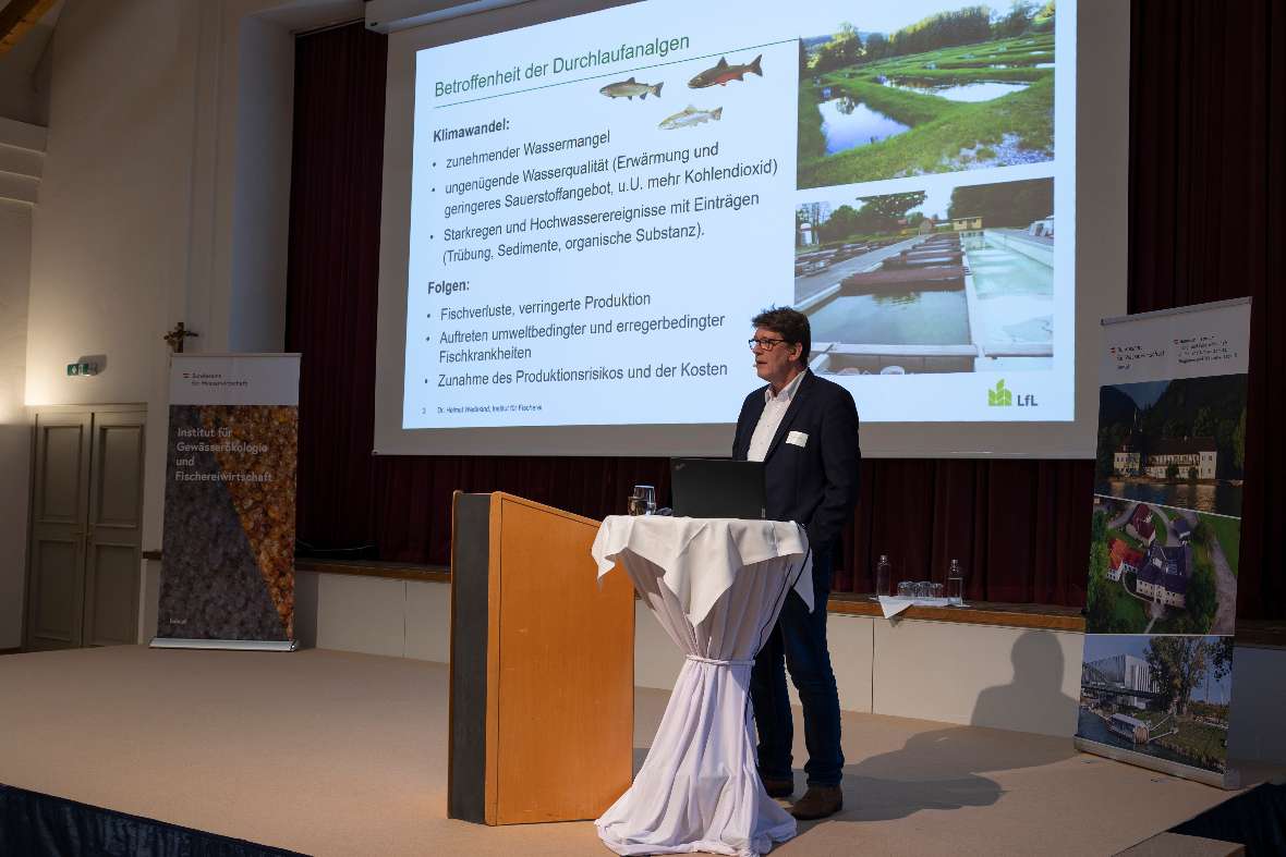 Helmut Wedekind (Bayerische Landesanstalt für Landwirtschaft, Institut für Fischerei Starnberg) steht an einem Stehpult vor einer Leinwand mit Präsentationsfolie über Klimawandel und Durchlaufanlagen in der Aquakultur.