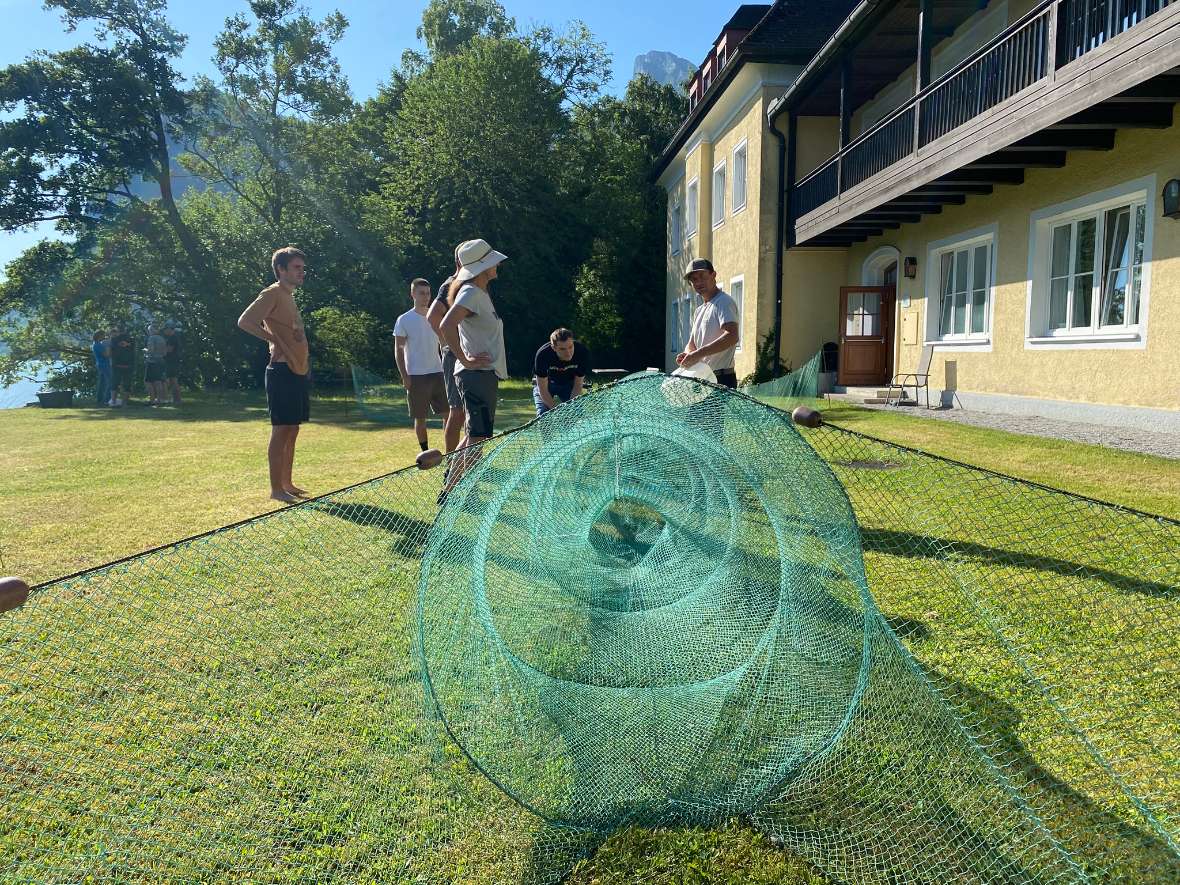 Gruppe von Personen auf einer Wiese, die eine große grüne Fischreuse ausbreiten und besprechen.