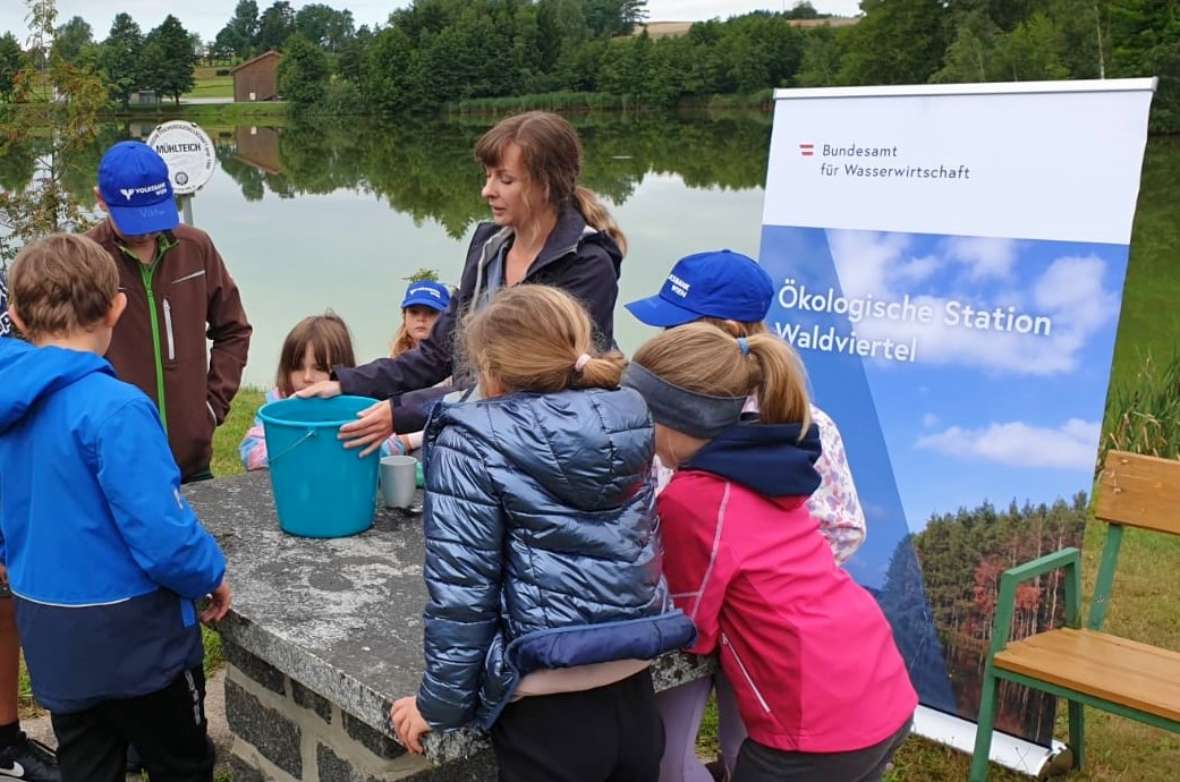 Gruppe von Kindern und BAW-Mitarbeiterin an einem Teich mit einem blauen Eimer und einem Banner der Ökologischen Station Waldviertel.
