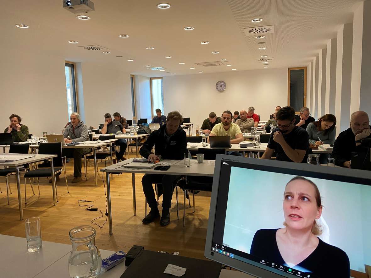 Vortragende des Institutes sowie externe lehrten sowohl in Präsenz als auch aus der Ferne. Man sieht die Kursteilnehmer:innen im Lehrsaal sitzen. Rechts im Bild ist ein Bildschirm mit einer Vortragenden. 