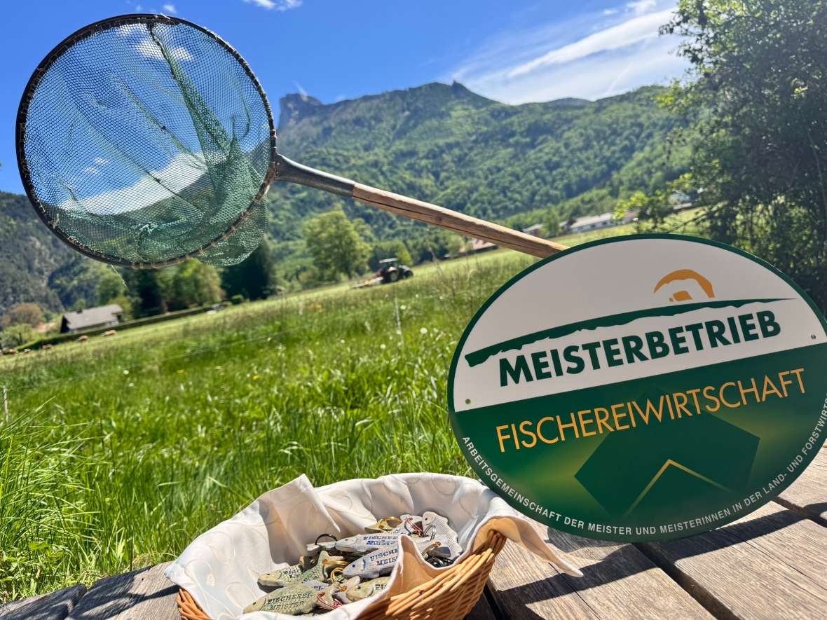 Fischkescher mit grünem Netz lehnt auf einer Wiese vor Bergkulisse neben einem Schild mit der Aufschrift MEISTERBETRIEB FISCHEREIWIRTSCHAFT.