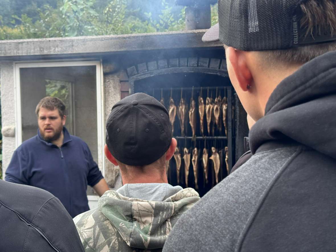Die Gruppe steht vor einem Räucherofen, in dem mehrere Fische zum Räuchern hängen. FM Vogl Alexander erläutert den Vorgang und gibt dazu nähere Informationen.