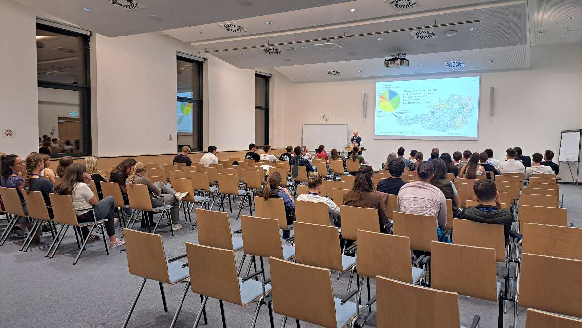 In einem Vortragssaal steht ein Mann an einem Pult, neben ihm ist eine große Leinwand auf der ein Bild aus dem Vortrag zu sehen ist. Im Saal sitzen ca. 50 Zuhörerinnen und Zuhörer.