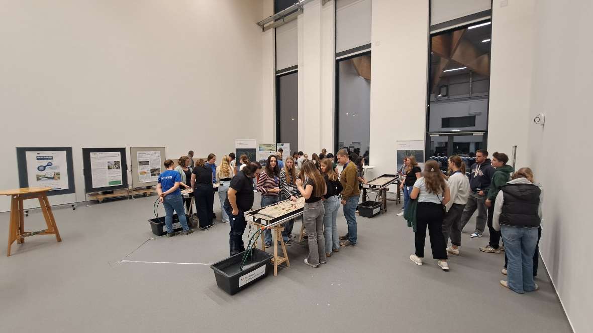 In einer großen Halle sieht man Schüler:innen an vier Experimentierrinnen stehen, die gerade mit verschiedenen Materialien Flüsse und Bäche in diesen Rinnen gestalten. Eine der Rinnen ist durch die vielen Personen verdeckt und daher nicht zu sehen.