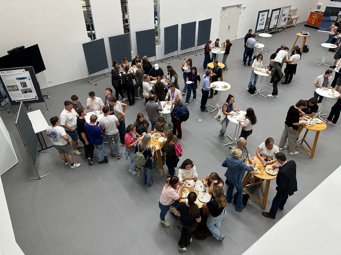 Blick von oben auf mehrere Stehtische in einem großen Raum. Zahlreiche Teilnehmerinnen und Teilnehmer essen Gemüse-Curry mit Reis.