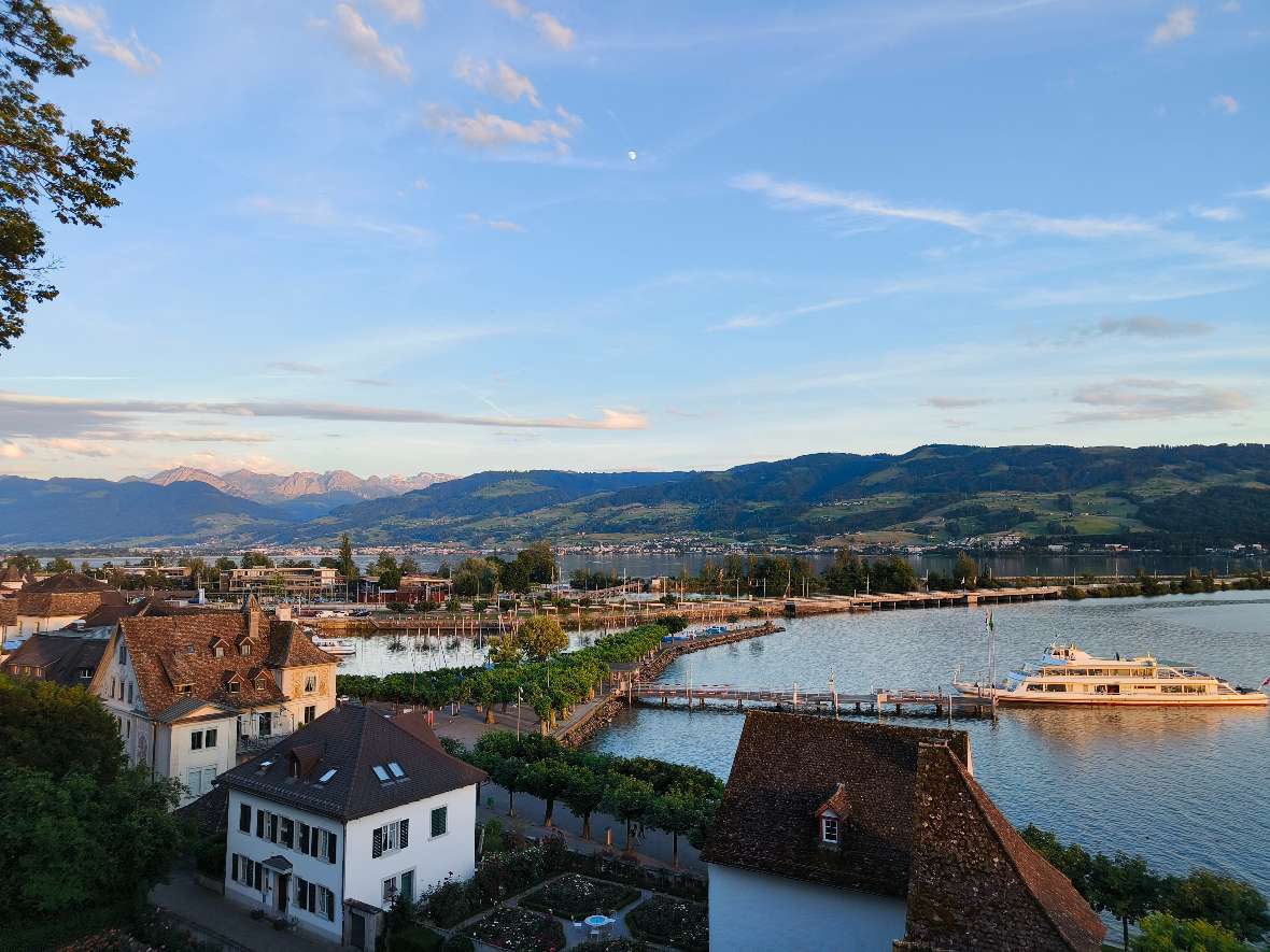 Ein Ausblick in der Abenddämmerung in Rapperswil auf den Hafen, den Zürichsee, die Landverbindung auf die andere Seite des Sees und die Berge im Hintergrund.
