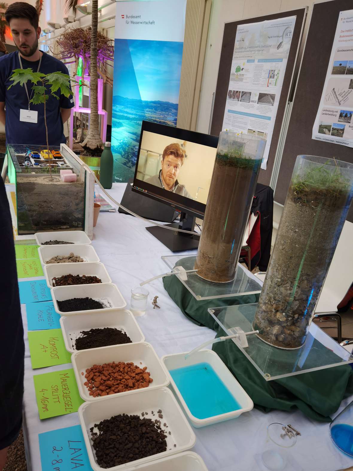 Man sieht die verschiedenen Baumsubstrate auf einem Tisch im Palmenhaus. Plakate und ein Bildschirm mit Videos sind im Hintergrund zu sehen.