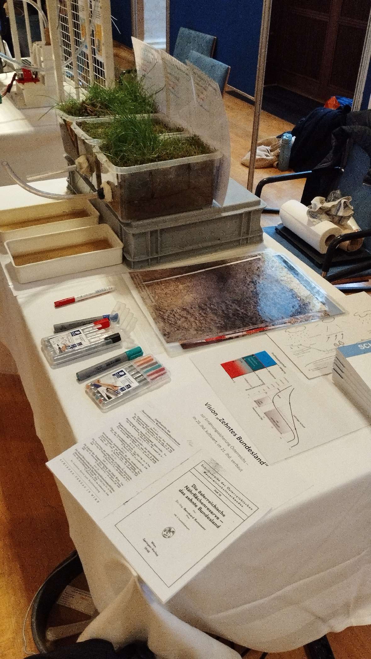 Tisch mit Gras in durchsichtigen Behältern, Stiften, Dokumenten und gedruckten Diagrammen auf weißer Tischdecke.