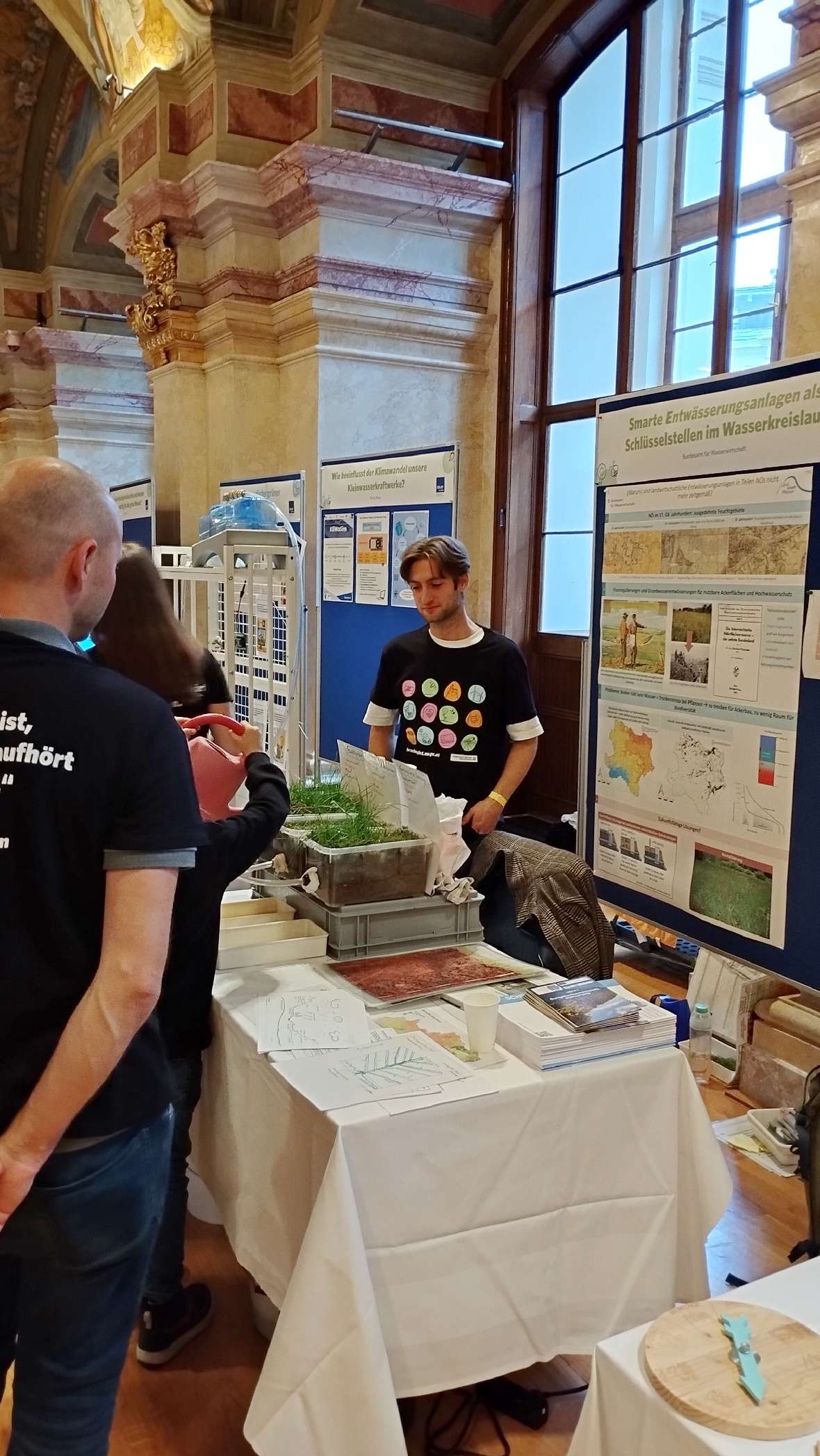 Messestand mit Informationsplakaten zu Starkregen und Wasserkreislauf, Tisch mit Gras und Flyern, Personen im Gespräch.