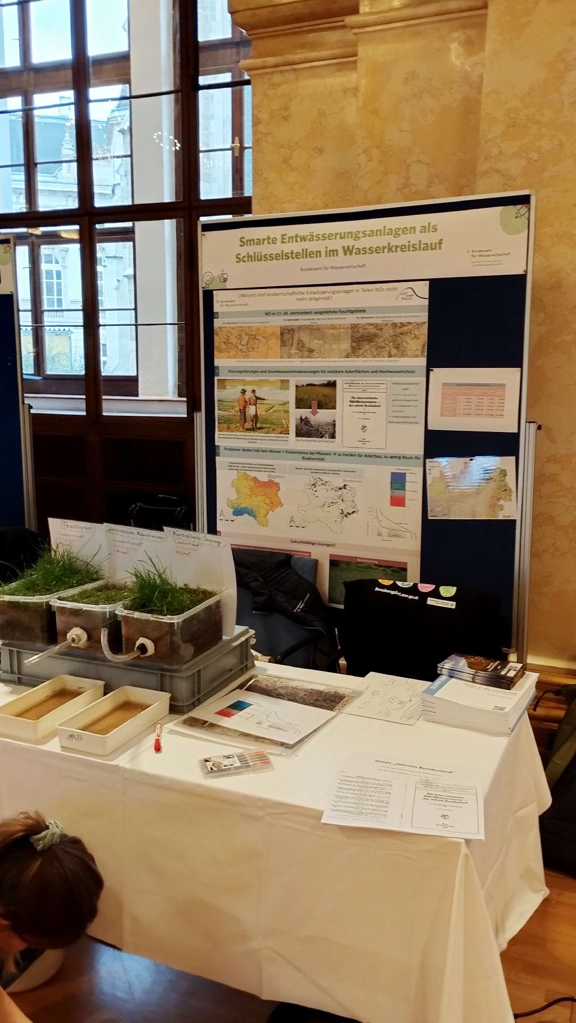 Messestand mit Poster 'Smarte Entwässerungsanlagen als Schlüsselstellen im Wasserkreislauf', Informationsmaterial und Pflanzen in rechteckigen Behältern auf Tisch.