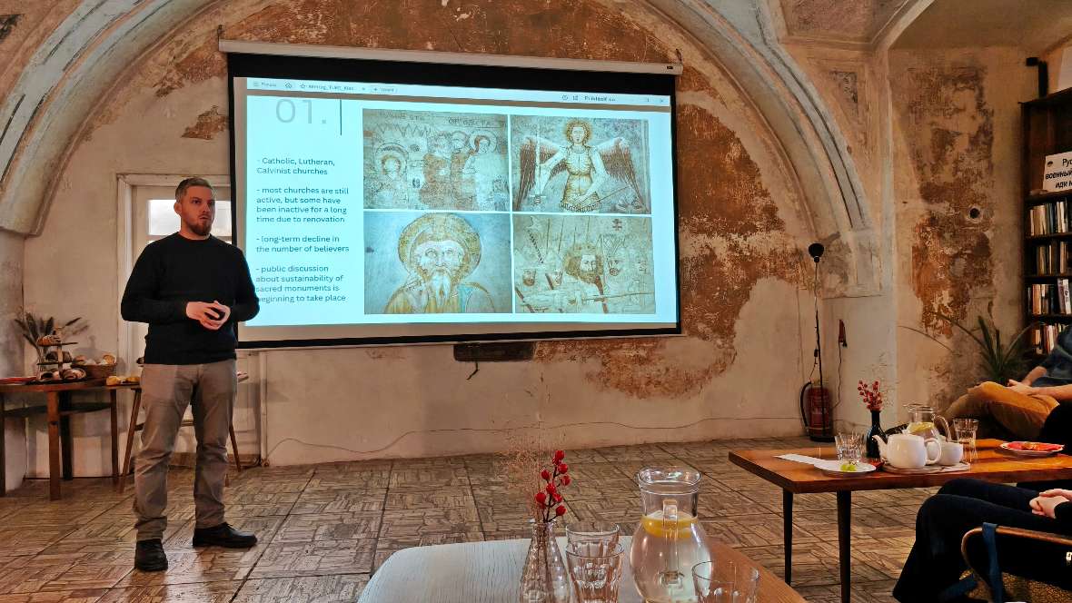 Im Kloster Rožňava wurden die Projektpartner über die Geschichte und Gefährdung des Klosters informiert. Person steht vor einer Leinwand mit einer Präsentation, die vier religiöse Fresken zeigt, in einem Raum mit gewölbter Decke und Steinboden.