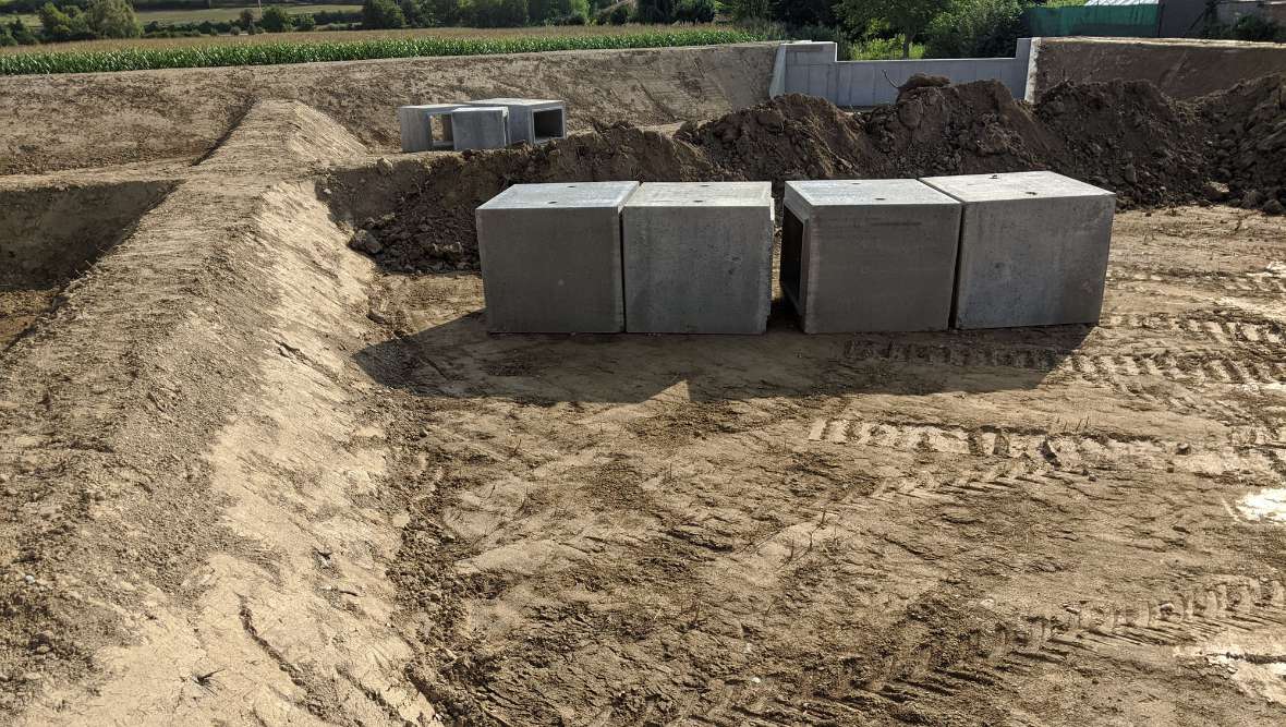 Das Bild zeigt einen der aus Erde aufgeschütteten Dämme und mehrere Betonfertigteile (Betonkästen), durch die das Wasser von einem Becken in das andere fließen kann. Im Hintergrund sieht man eine Betonmauer, die den Auslauf enthält
