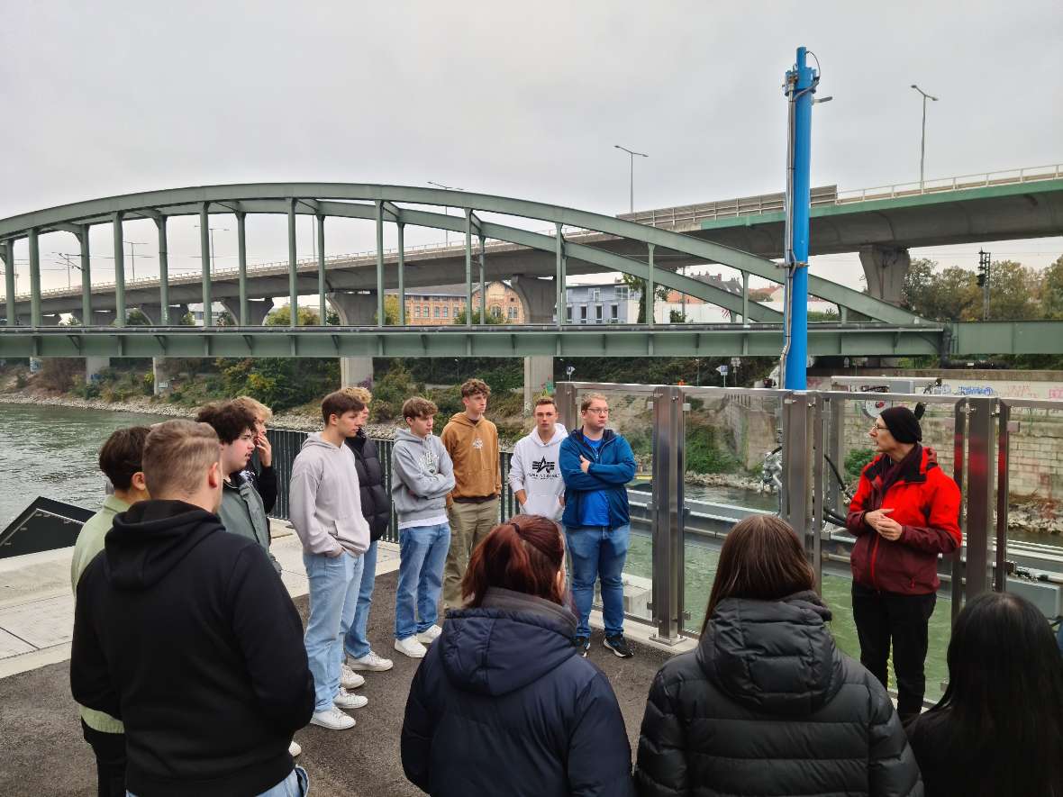 Eine Gruppe von Schülerinnen und Schülers steht am Ufer des Donaukanals. Im Hintergrund sieht man eine Eisenbahnbrücke, eine Straßenbrücke und einige Häuser.