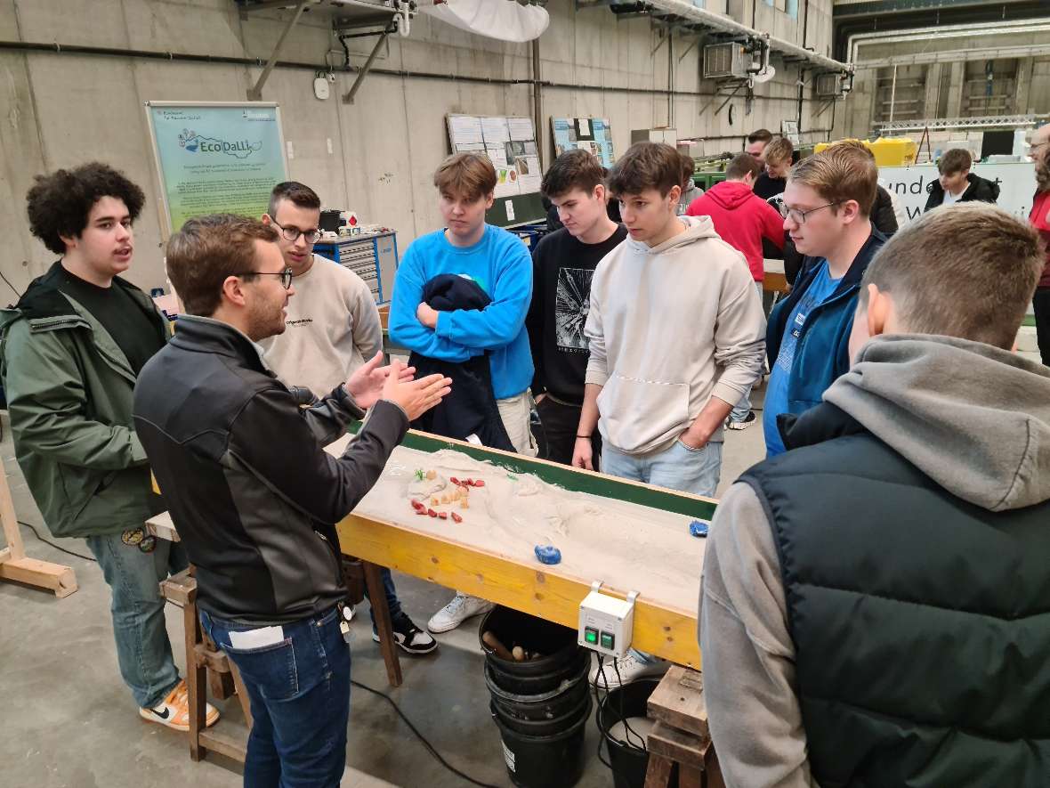 Man sieht die Morphologie-Rinne unseres Instituts, deren Funktion einer Gruppe von Schülern erläutert wird. Diese Rinne besteht aus einer mit Holz gebauten Wanne, die mit Sand gefüllt ist. Wenn durch die Wanne Wasser läuft, bildet sich im Sand ein Flussbett. Häusern, Bäumen und bunten Steinen können die Landschaft und der Flusslauf gestaltet werden.