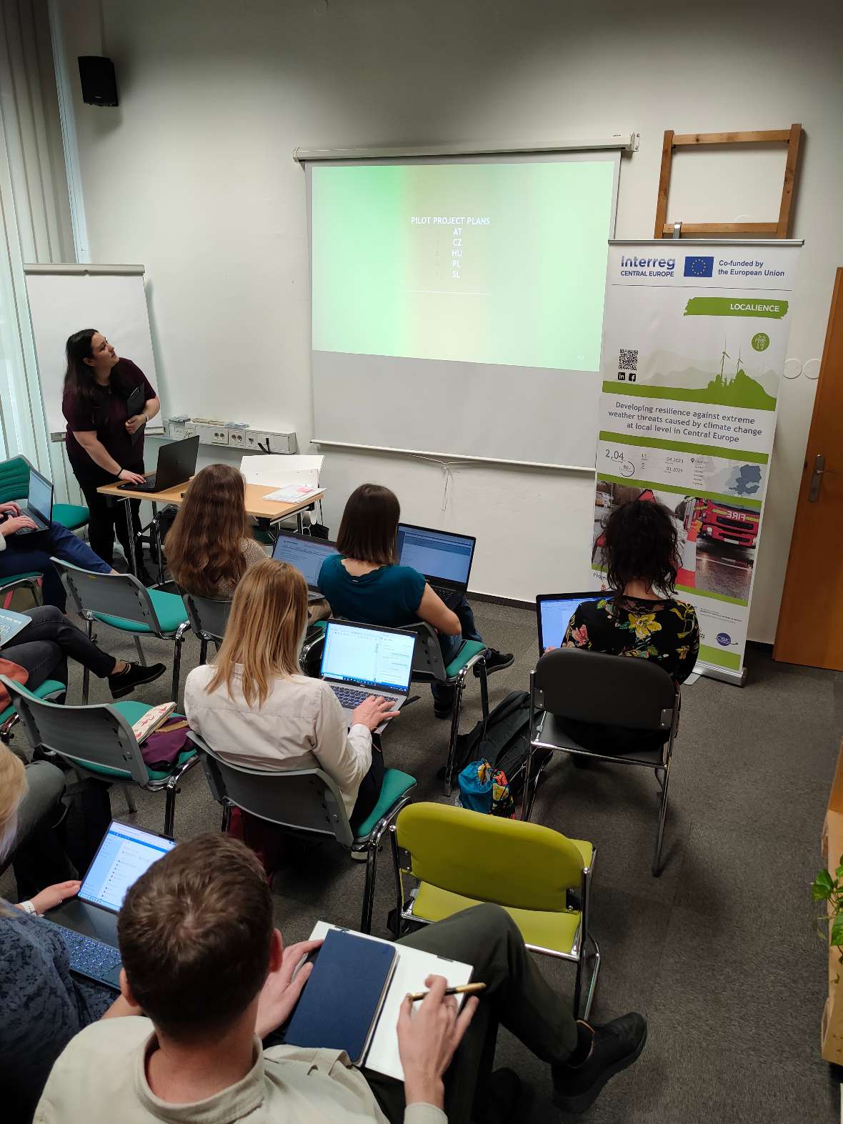 Die Projektteilnehmer:innen sitzen mit ihren Laptops vor einer Leinwand auf der eine PowerPoint publiziert ist.