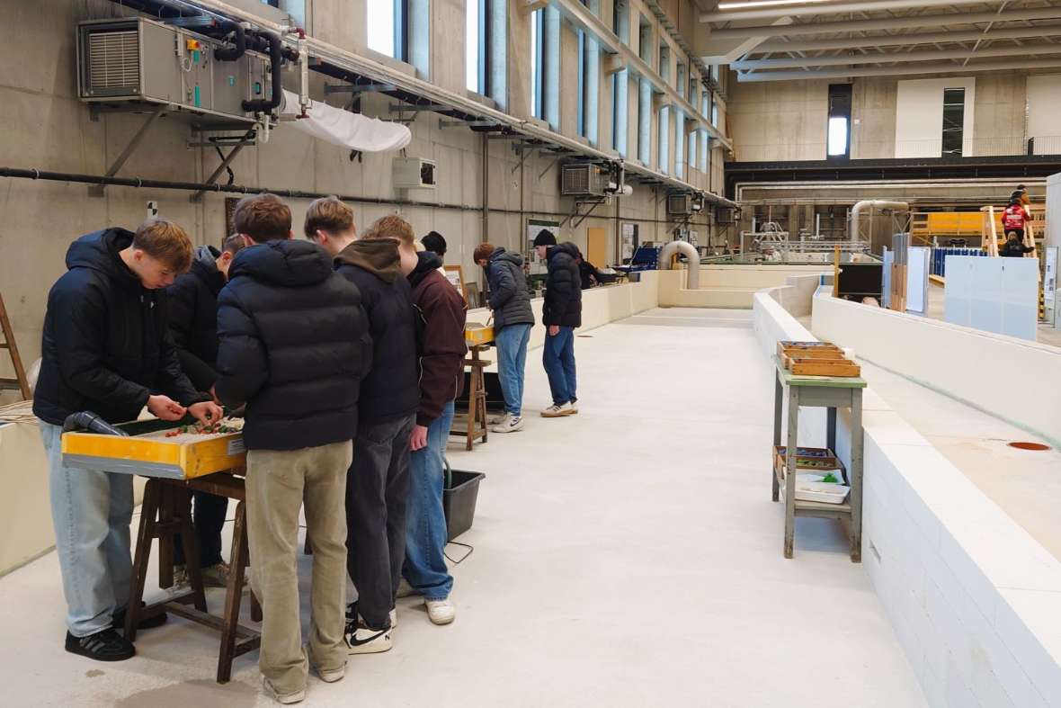 In einer großen Laborhalle sieht man Schüler an zwei Experimentierrinnen stehen, die gerade mit verschiedenen Materialien in diesen Experimentierrinnen bauen.