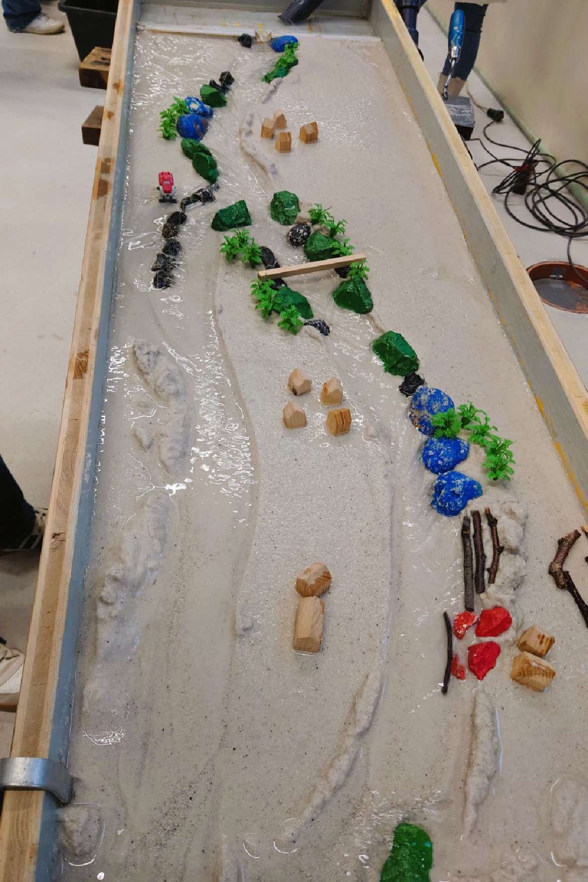 Vogelperspektive einer mit Sand gefüllten Experimentierrinne, in der Steine und Hölzer das durch mehrere Krümmungen fließende Wasser lenken.