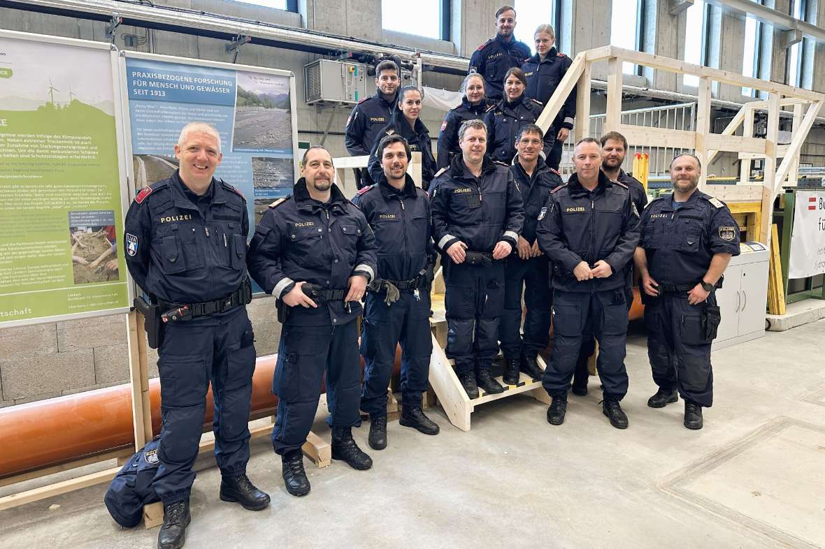 Blindenbeschreibung:  Ein Gruppenbild mit 14 Polizistinnen und Polizisten, die auf einer Treppe aus Holz im Wasserbaulabor stehen.