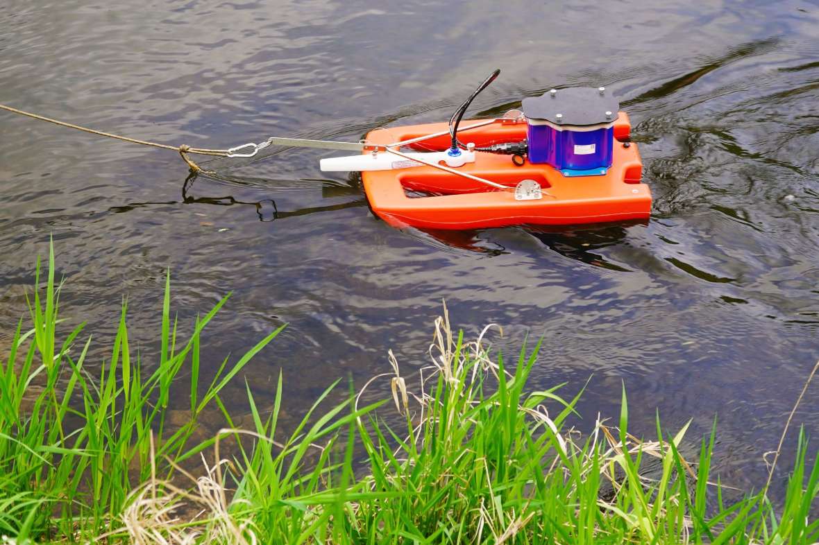 Ein rotes Messboot schwimmt in Ufernähe mit einem blauen akustischen Messgerät (ADCP).