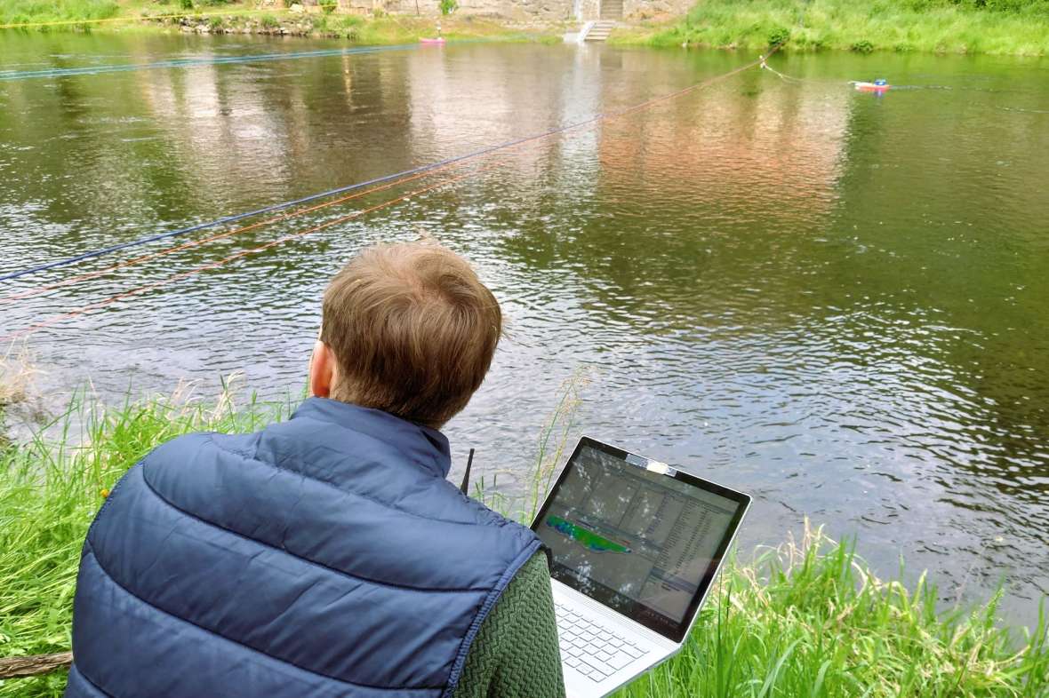 Blick über die Schulter eines Kollegen auf den Fluss, auf dem ein Messboot über den Querschnitt gezogen wird. Am Laptop werden die Messergebnisse verfolgt.