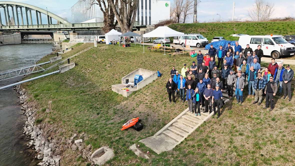 Zu sehen ist eine Gruppe von etwa 50 Personen, die an einer Böschung stehen, die zum Donaukanal hinabführt. Auf der Böschung liegen Messgeräte, im Hintergrund sind Veranstaltungszelte und die Autos der Teilnehmenden zu sehen. Ganz im Hintergrund ist eine Eisenbrücke über den Donaukanal und ein Teil des Wasserbaulabors zu erkennen.