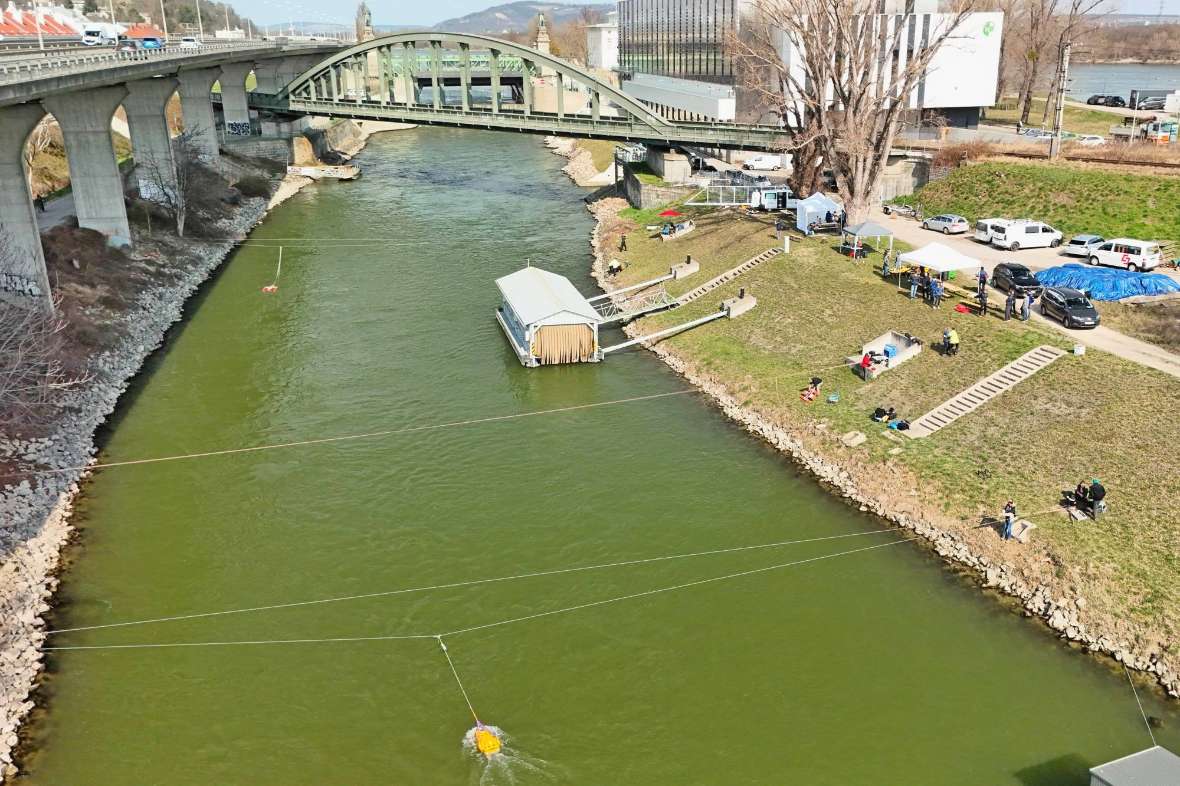 Eine Drohnenaufnahme von oben mit einem Überblick über den Donaukanalabschnitt, über den drei Seile gespannt sind, an denen wiederum Messboote fixiert sind. In Blickrichtung am rechten Ufer sind Messteams, Zelte und Autos der Teilnehmenden zu erkennen. Im Hintergrund führt eine Eisenbahnbrücke über den Donaukanal, und Teile des Wasserbaulabors sind zu erkennen.