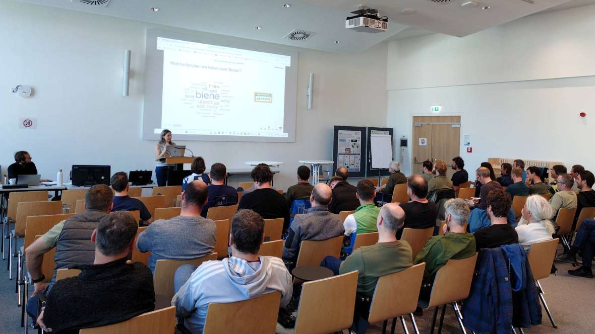 Ein großer Hörsaal mit Einzelbestuhlung, in dem ca. 50 Personen in Richtung Leinwand und Vortragende schauen. Die Vortragende steht am Rednerpult, auf der Leinwand ist eine Folie projiziert, die aber nicht genau erkennbar ist.