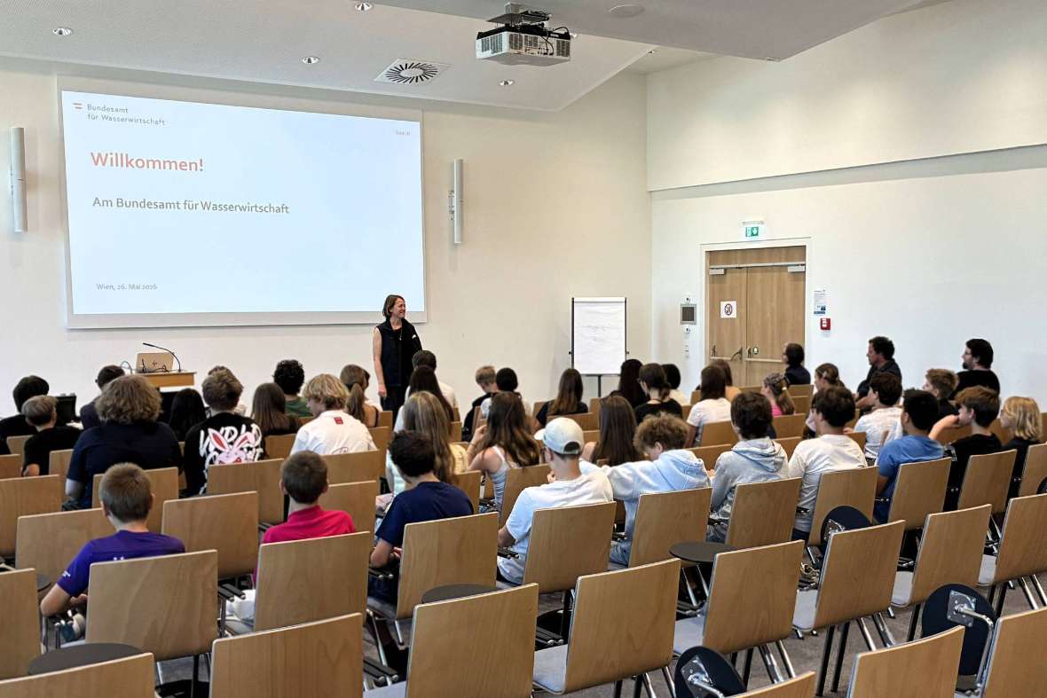 Ein großer Hörsaal mit Einzelbestuhlung, in dem ca. 40 Schüler und Schülerinnen in Richtung Leinwand und Vortragende schauen. Die Vortragende steht vor der Leinwand, auf dieser ist eine Willkommens-Folie projiziert.
