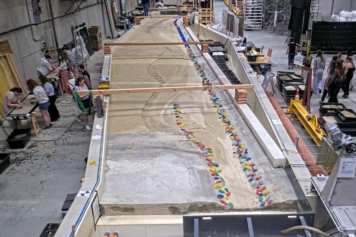 Vogelperspektive auf einen großen Modellversuch, der einen geschwungenen Flusslauf enthält, in der Laborhalle. Rechts davon ist eine Kleingruppenführung zu sehen, links vom Modellversuch stehen die Experimentierrinnen, an denen eine Besuchergruppe arbeitet.