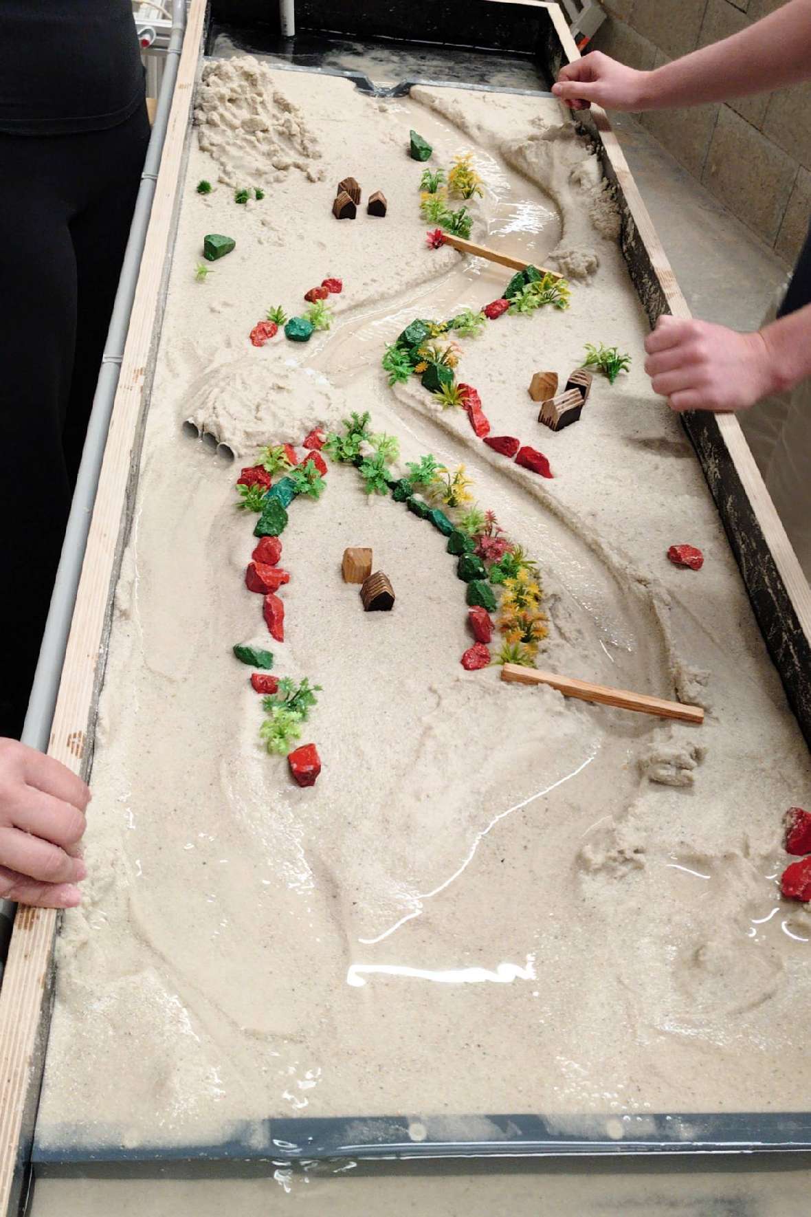 Vogelperspektive einer mit Sand gefüllten Experimentierrinne, in der Steine und Hölzer das durch mehrere Krümmungen fließende Wasser lenken.