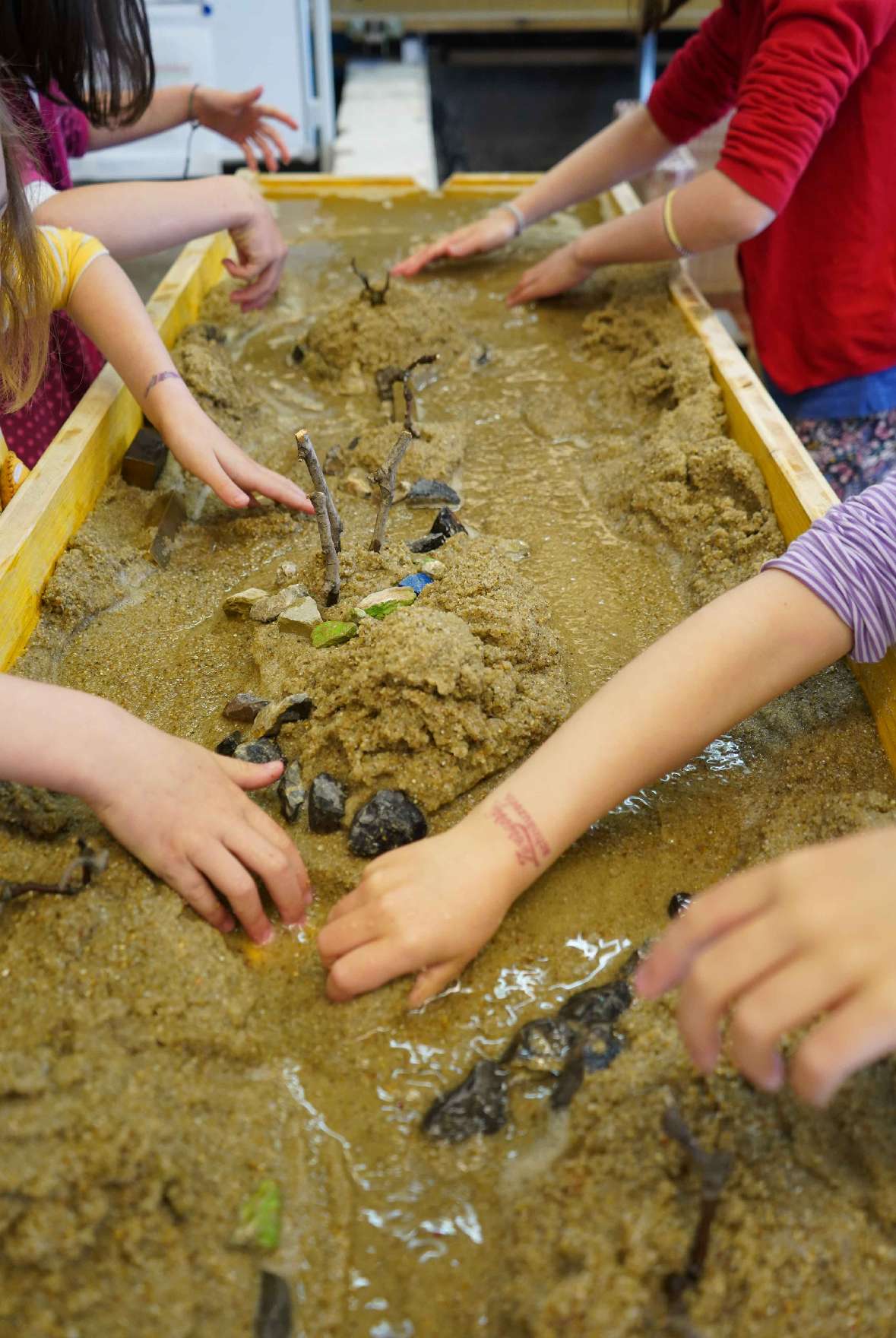 : In einer etwa zwei Meter langen Rinne befindet sich Sand, durch den Wasser fließt. Acht Hände und das Wasser arbeiten daran, eine Flusslandschaft in dem Sand zu gestalten.