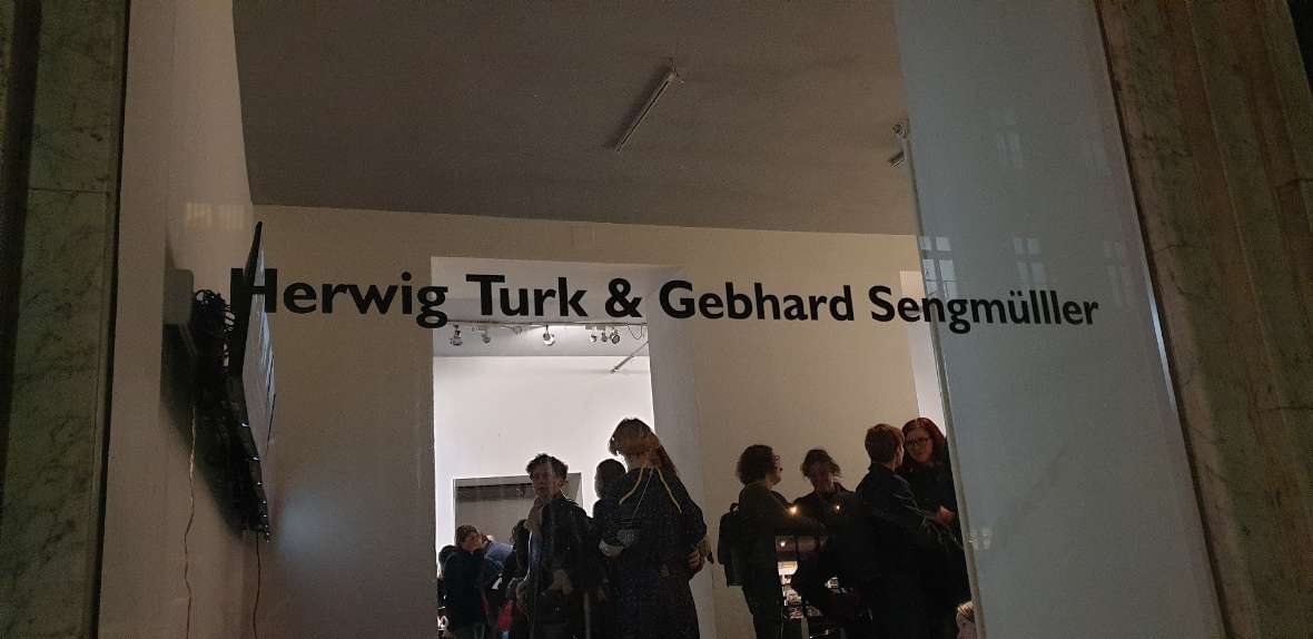 Das Bild besteht aus zwei Teilen. In beiden Teilen sieht man von der Straße durch die Schaufenster in die Galerie. Im linken Bild sieht man viele Menschen bei der Ausstellungseröffnung und die Namen der Künstler Herwig Turk und Gebhard Sengmüller am Glas geschrieben. Am rechten Bild steht der Name der Ausstellung am Glas des Schaufensters. Direkt hinter dem Fenster steht ein Kunstwerk (ein großer Mann mit langem Bart, hergestellt aus gelbem Schaumstoff).