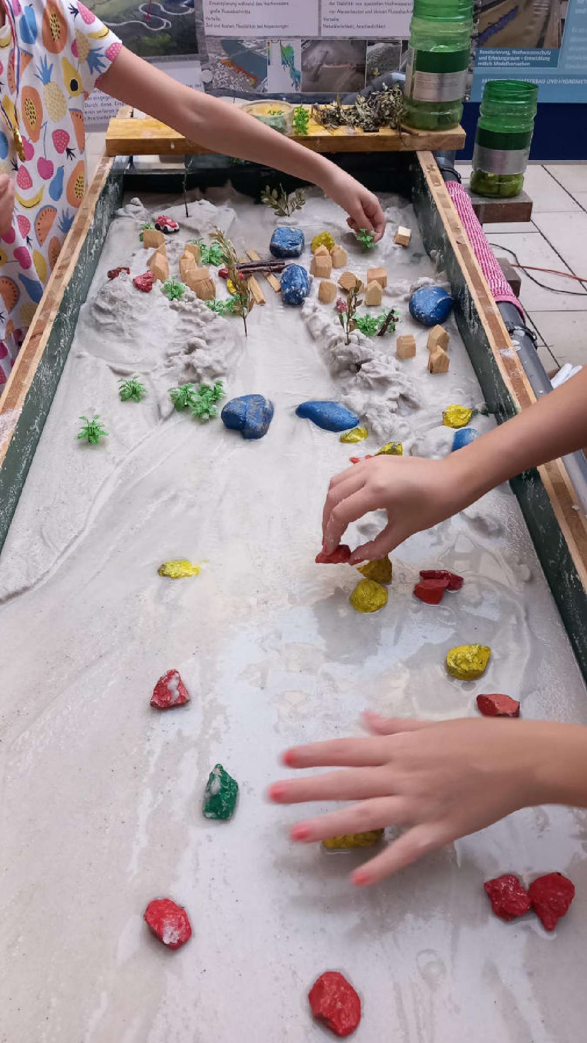 Das Bild zeigt die circa 1 Meter breite Rinne unseres Modellflusses, die mit feinem Sand gefüllt ist. Kinder stehen links und rechts vom Modellfluss und bauen mit bunten Steinen ihren eigenen Fluss.