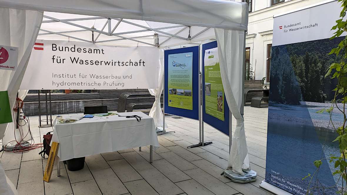 Das Foto zeigt die rechte Seite unseres Informationsstands auf einer Terrasse im Freien. Vorne steht ein Tisch mit Informationsmaterial, rechts davon 2 große Tafeln mit Postern zu unseren Projekten. Tisch und Poster sind durch ein Partyzelt geschützt.