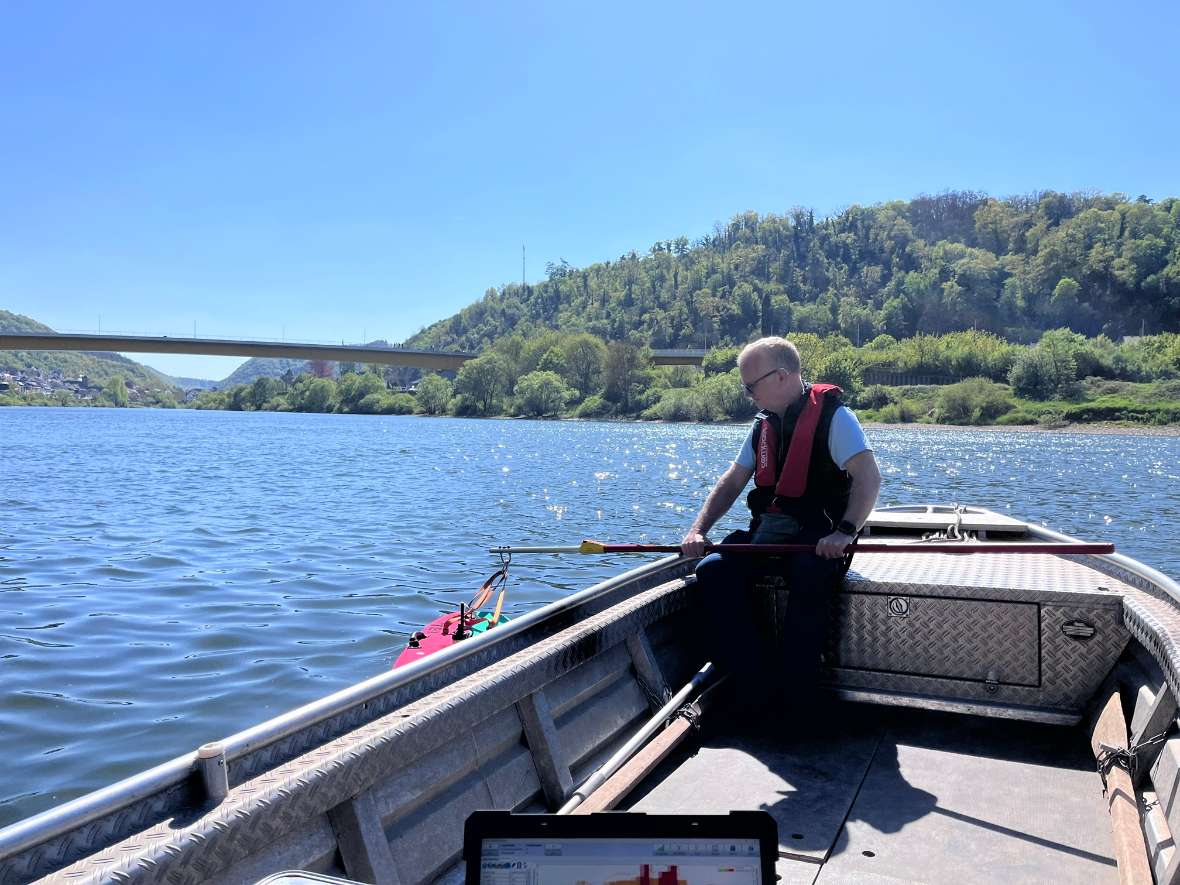 Herr Urbanek sitzt in einem Boot auf dem Fluss Mosel. Er hält eine Stange fest. Am Ende der Stange ist ein kleines Mess-Boot befestigt, das über das Wasser gleitet. Im Vordergrund steht ein Laptop, der die Daten speichert. Im Hintergrund sieht man das Ufer.