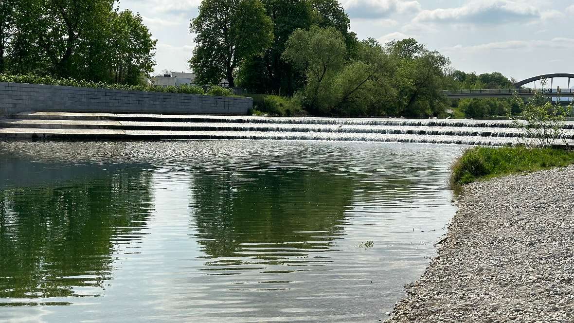 Der Blick geht gegen die Fließrichtung. Man sieht das Wasser über die 3 Stufen fließen. Im Hintergrund ist eine Brücke.
