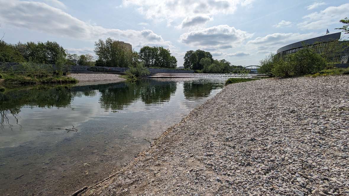 Das Bild zeigt einen Überblick mit dem langsam fließenden Fluss im Vordergrund, links und rechts sind große Kiesbänke. Im Hintergrund kommen die Sohlstufe und eine Brücke. Links steht ein Hochhaus und rechts ein Gebäude der niederösterreichischen Landesregierung.