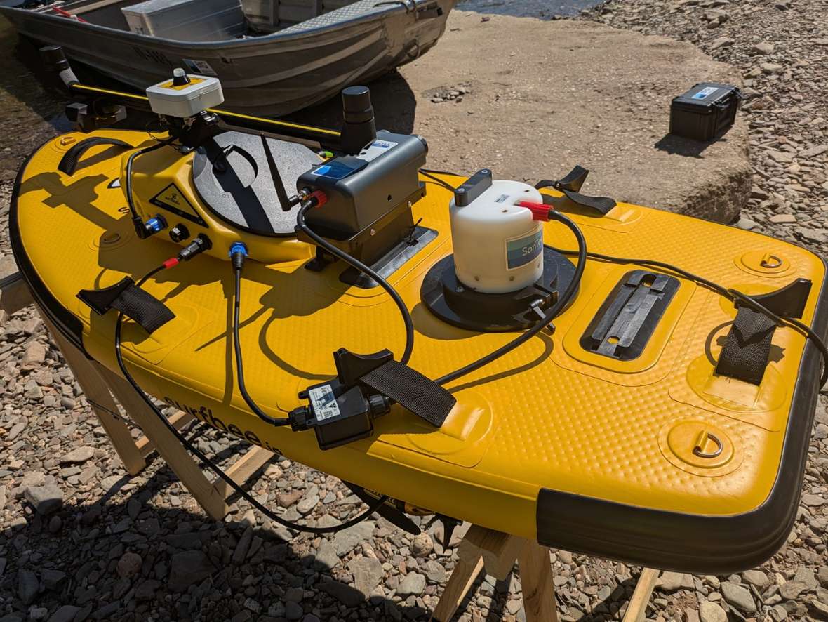 Seitliche Draufsicht auf ein gelbes ferngesteuertes ADCP-Messboot auf einem Holzstativ am Ufer. Mittig ist der ADCP-Sensor befestigt, weiter vorne befinden sich Akkuteile, GPS-Empfänger und Steuerungselemente für den Antrieb.