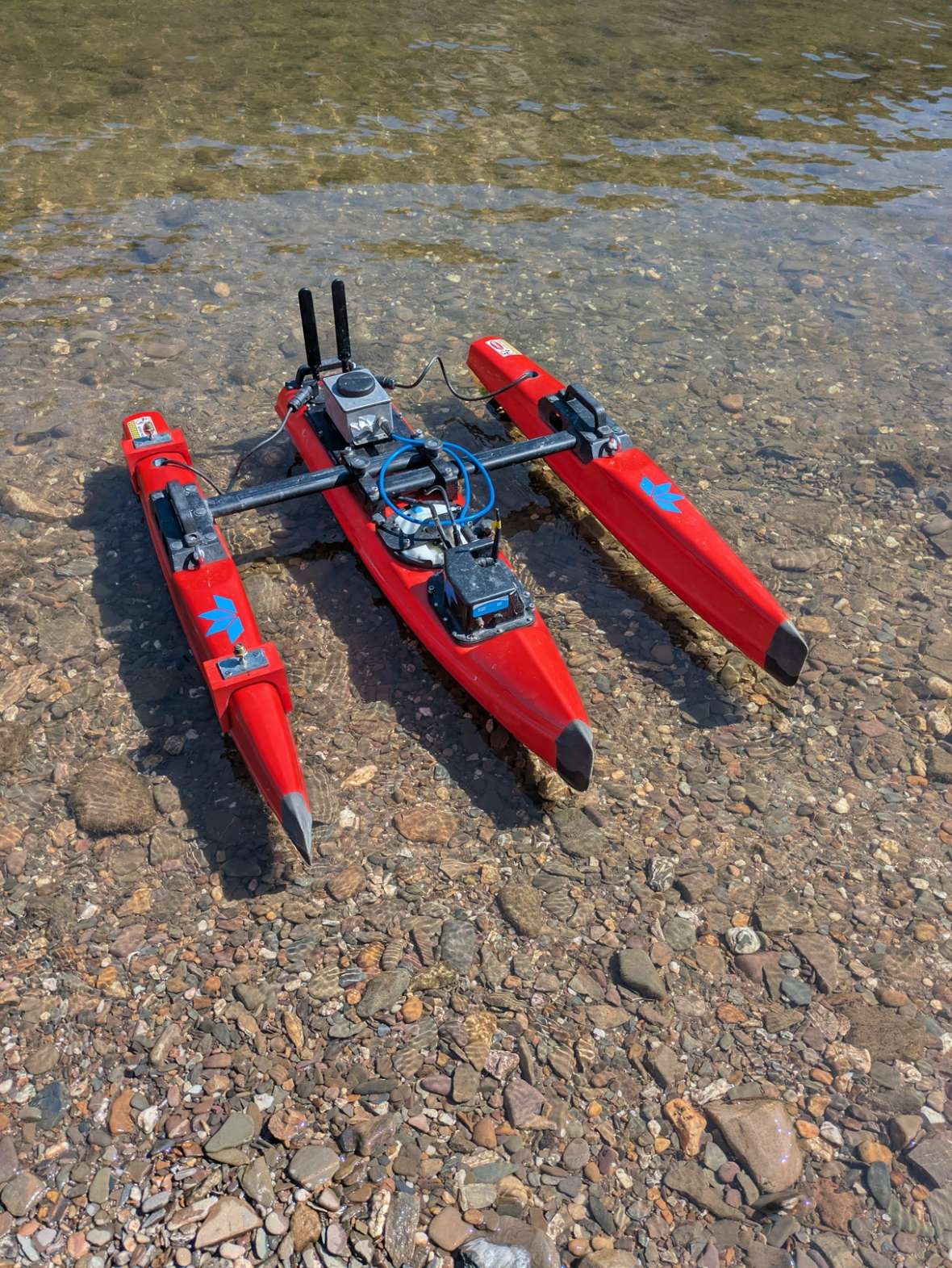 Man sieht ein ferngesteuertes rotes ADCP-Messboot von vorne, das ufernah im Wasser geparkt ist. Es handelt sich um einen Trimaran, dessen drei längliche Schwimmkörper auf der Oberseite mit einer Stange verbunden sind. Im mittleren Schwimmkörper sind der ADCP-Sensor, Funkteile, und der Akku befestigt.