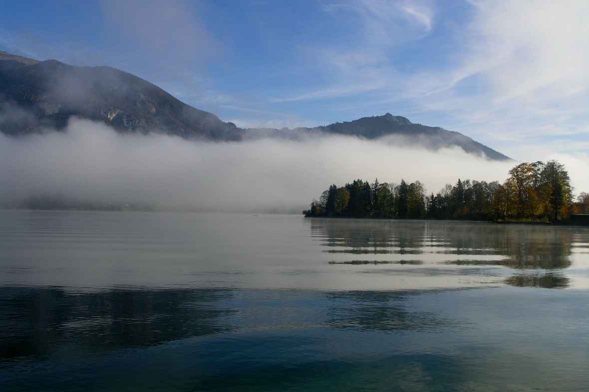 Photo directly over water, trees on the right. Behind it mountain, in front of it a dense cloud.