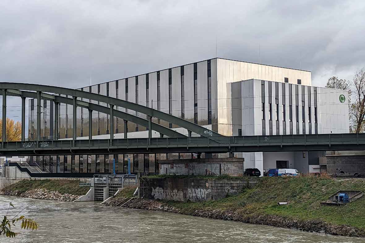 Das Bild zeigt, vom Süden aus schräg aufgenommen den Donaukanal im Vordergrund und das Gebäude des Wasserbaulabors am anderen Ufer. Es ist ein sehr helles Gebäude. Die Fenster sind als vertikale, dunklere Streifen zu erkennen. Vor dem Gebäude ist eine Eisenbahnbrücke sichtbar, die über den Donaukanal verläuft. Bei der Anlieferung zum Gebäude sind einige Firmenwagen zu sehen. Die Aufnahme erfolgte bei herbstlichen und wolkigem Wetter.