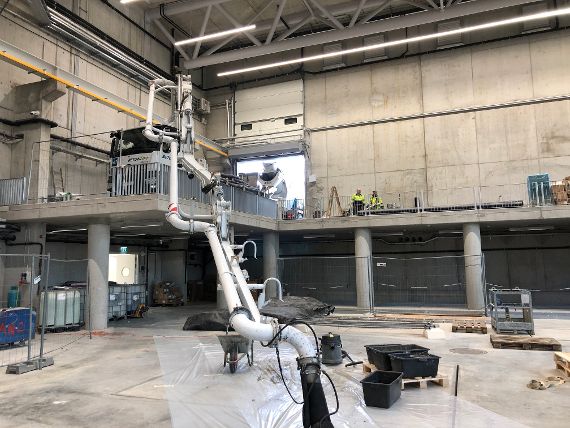 The picture shows a large hall with a platform standing on columns approx. 3 m above the hall floor. A truck with a concrete pump is standing on this platform. A pipe leads to the hall floor to transport the concrete to the placement site.