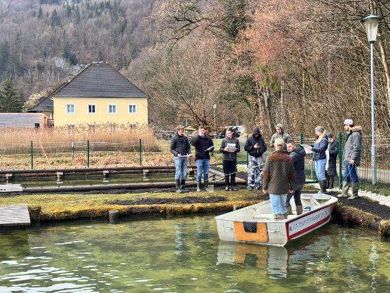 The participants are standing by the institute pond in rubber boots. There is a boat in the pond. The participants are recording hydrological data directly at the pond.