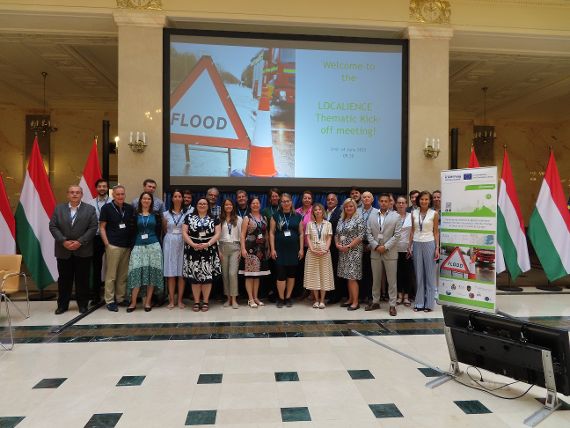 On the group photo in the Marble Hall of the Hungarian Ministry of Interior, 26 people are standing in front of a large presentation wall. The wall displays the text "Welcome to the Localience Thematic Kick off Meeting!". Hungarian flags can be seen on the left and right sides of the individuals.