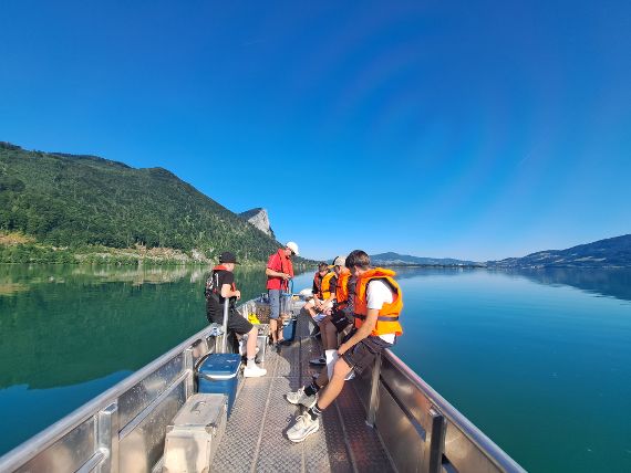 Sechs Personen mit orangen Schwimmwesten auf einem Boot bei Seenprobenahme auf dem Mondsee vor bewaldeten Bergen und klarem Himmel